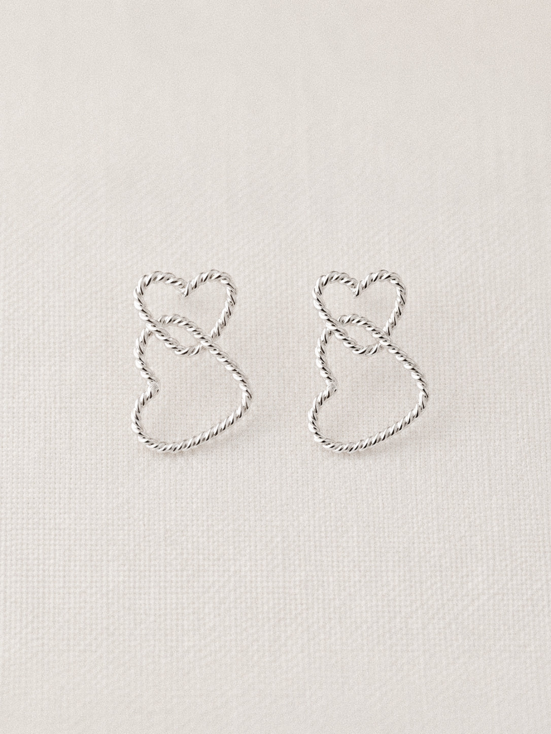 Sterling silver twisted wire double heart stud earrings with two interlocking rope-textured hearts on a soft linen background